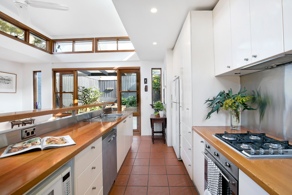 This is an example of a mid-sized transitional single-wall open plan kitchen in Sydney with a single-bowl sink, shaker cabinets, white cabinets, wood benchtops, stainless steel appliances, terra-cotta floors, with island and brown benchtop.