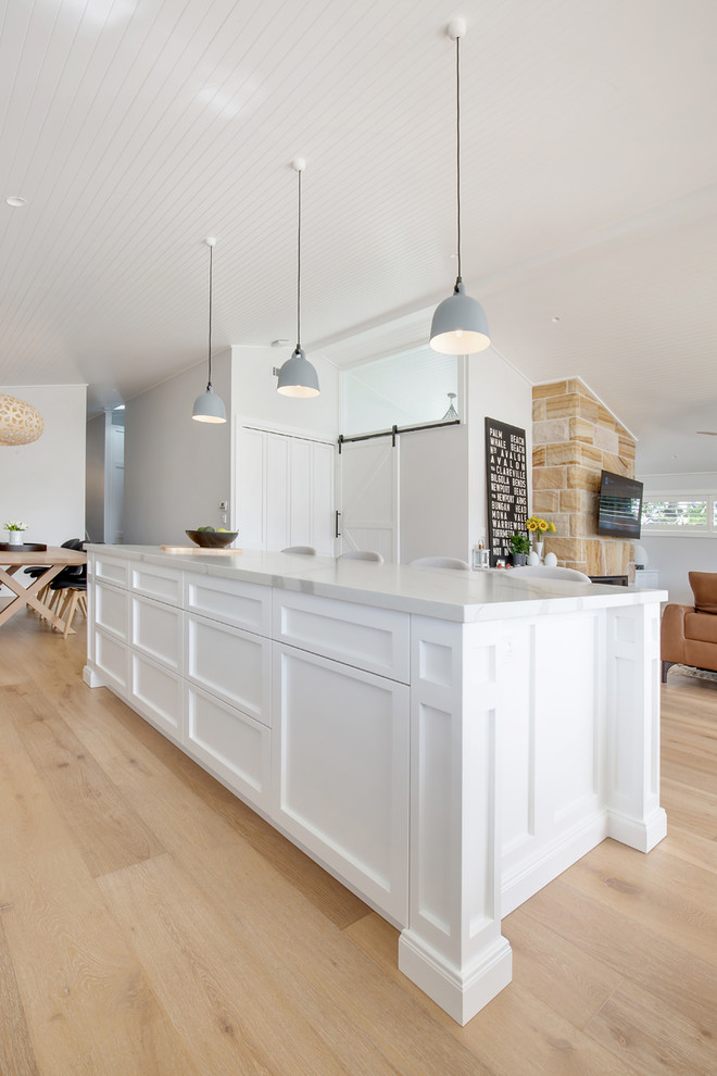 Mid-sized beach style l-shaped open plan kitchen in Sydney with shaker cabinets, white cabinets, quartz benchtops, grey splashback, medium hardwood floors, with island, brown floor and white benchtop.