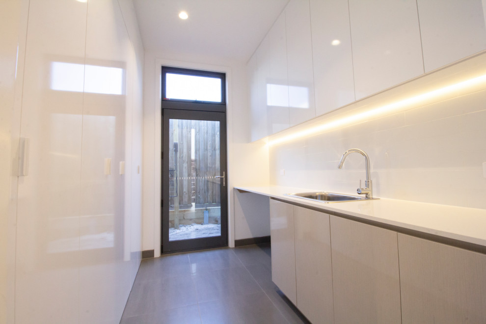 This is an example of a mid-sized contemporary galley dedicated laundry room in Melbourne with a single-bowl sink, recessed-panel cabinets, white cabinets, concrete benchtops, white walls, ceramic floors, a side-by-side washer and dryer, grey floor and white benchtop.