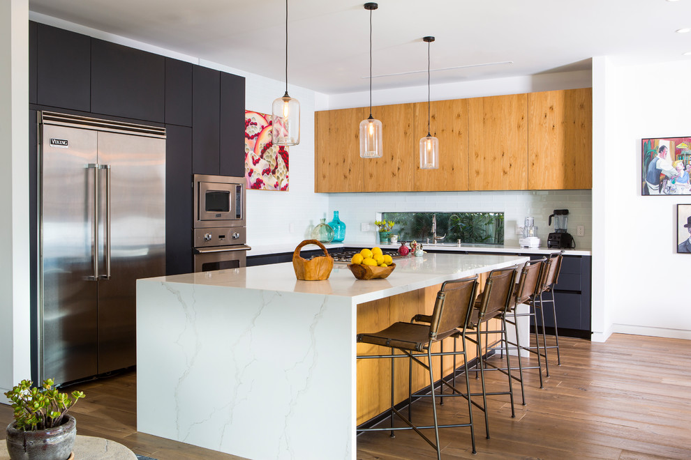 Photo of a large contemporary l-shaped kitchen in Los Angeles with an undermount sink, flat-panel cabinets, solid surface benchtops, white splashback, subway tile splashback, stainless steel appliances, medium hardwood floors, with island, brown floor, white benchtop and grey cabinets.