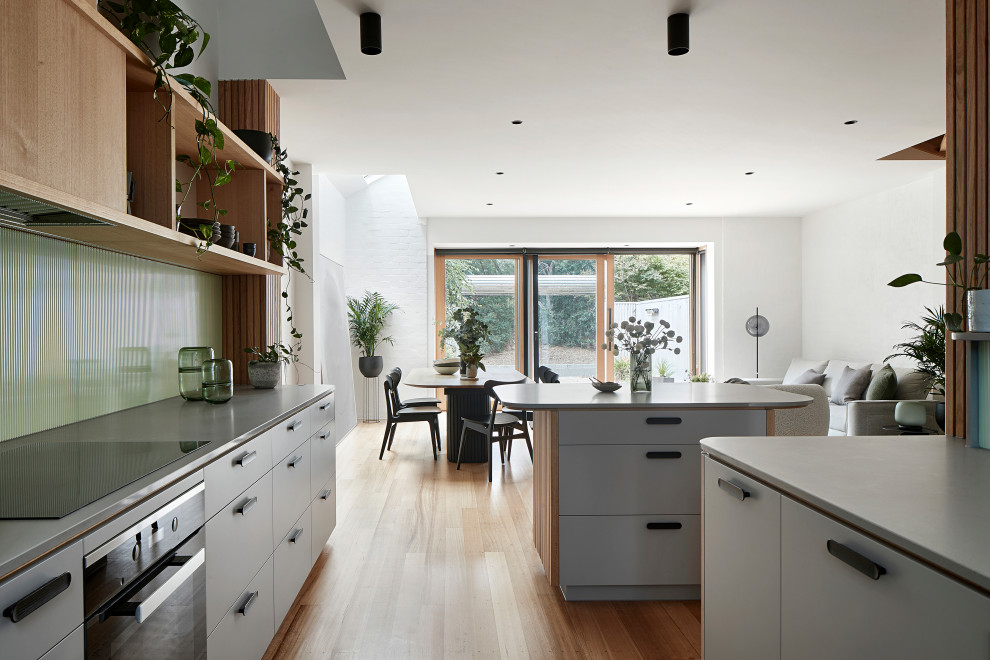 Photo of a mid-sized modern u-shaped open plan kitchen in Melbourne with an undermount sink, flat-panel cabinets, grey cabinets, quartz benchtops, green splashback, glass sheet splashback, black appliances, light hardwood floors, with island and grey benchtop.
