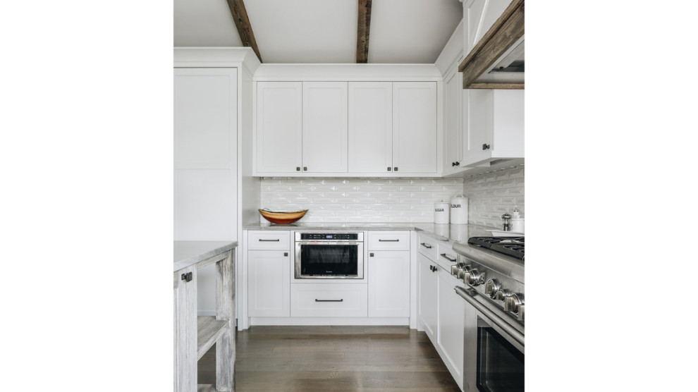 Diseño de cocina de estilo de casa de campo con puertas de armario blancas, salpicadero blanco, salpicadero de azulejos tipo metro, electrodomésticos de acero inoxidable, suelo de madera clara y vigas vistas
