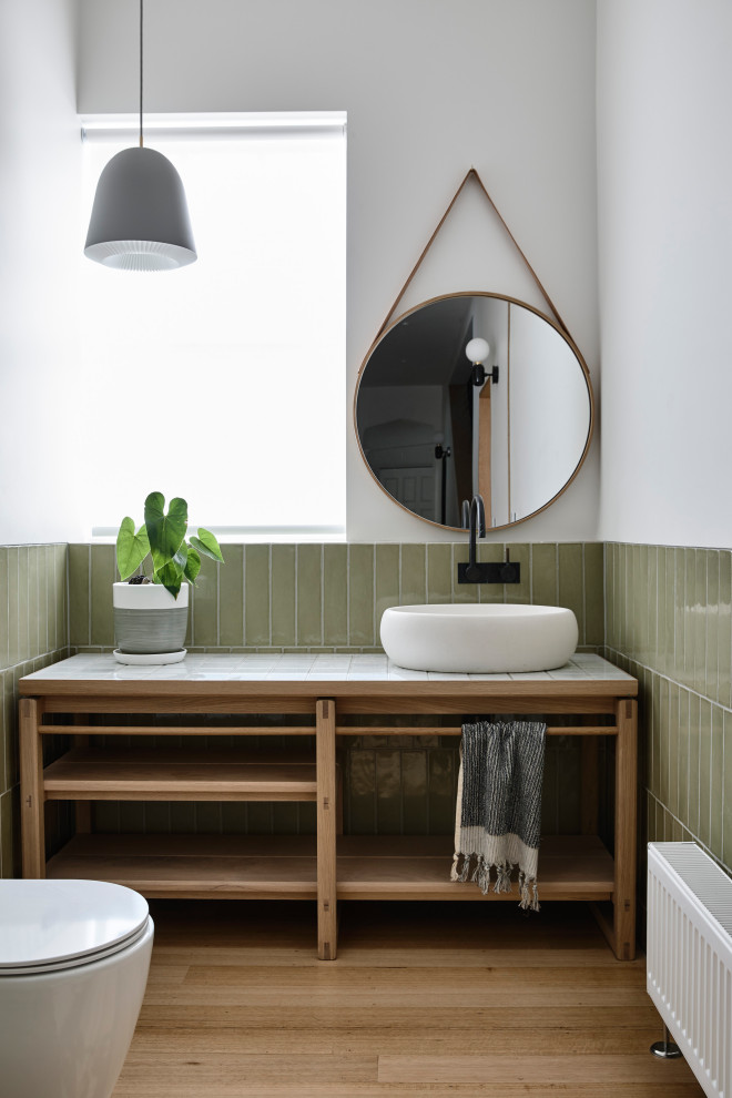 This is an example of a mid-sized contemporary powder room in Melbourne with furniture-like cabinets, light wood cabinets, a wall-mount toilet, green tile, ceramic tile, white walls, light hardwood floors, a vessel sink, tile benchtops, brown floor and white benchtops.