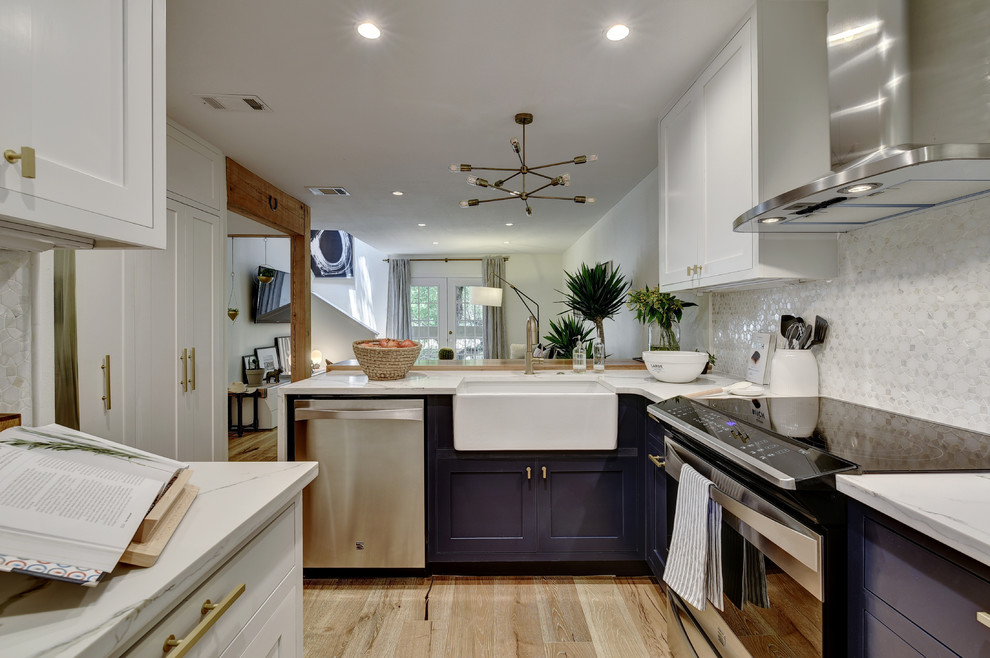 Photo of a transitional l-shaped open plan kitchen in Austin with a farmhouse sink, shaker cabinets, blue cabinets, marble benchtops, white splashback, stainless steel appliances and light hardwood floors.