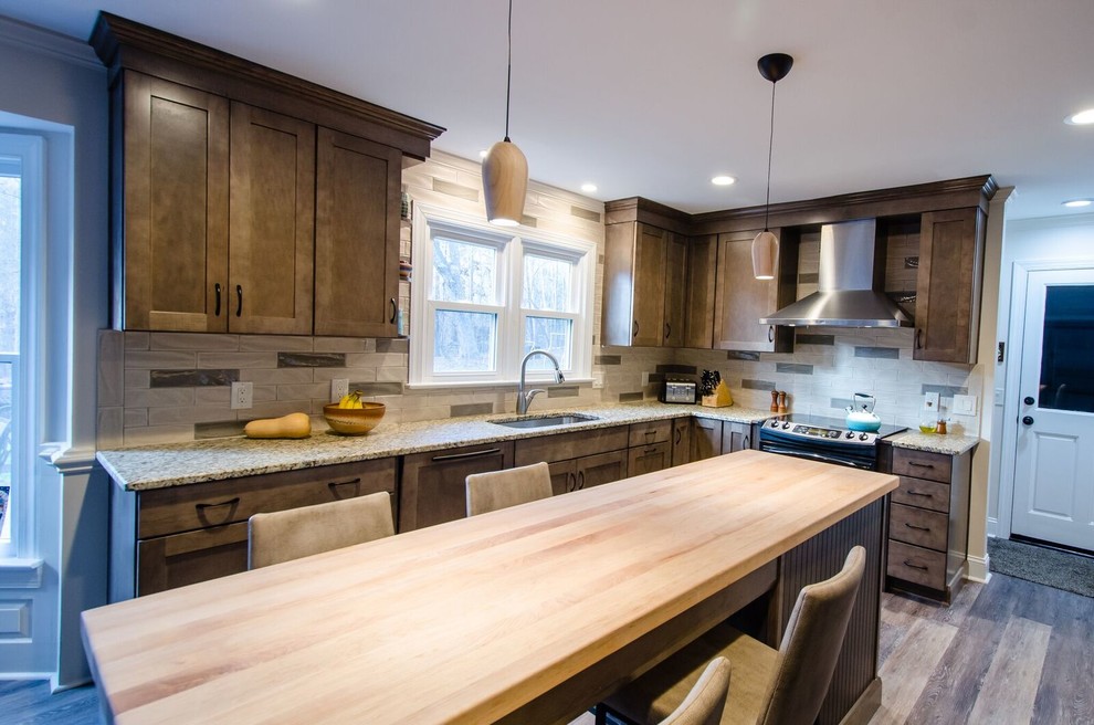Inspiration for a mid-sized arts and crafts galley open plan kitchen in Raleigh with an undermount sink, recessed-panel cabinets, brown cabinets, granite benchtops, beige splashback, stone slab splashback, stainless steel appliances, light hardwood floors and with island.