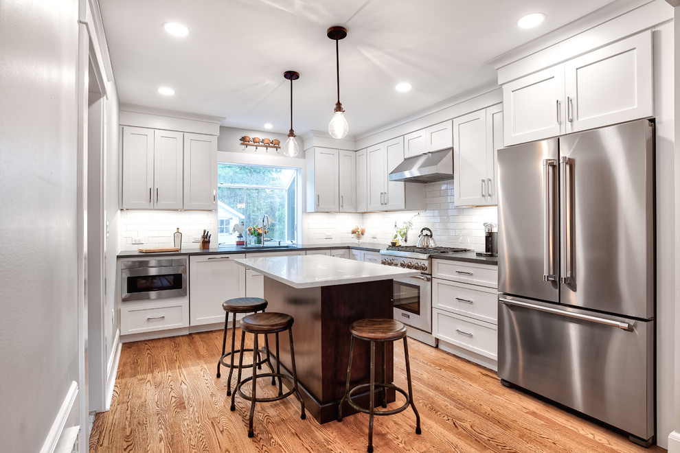 This is an example of a mid-sized country l-shaped kitchen in New York with white cabinets, white splashback, subway tile splashback, stainless steel appliances, with island, brown floor, black benchtop, an undermount sink, shaker cabinets, soapstone benchtops and medium hardwood floors.