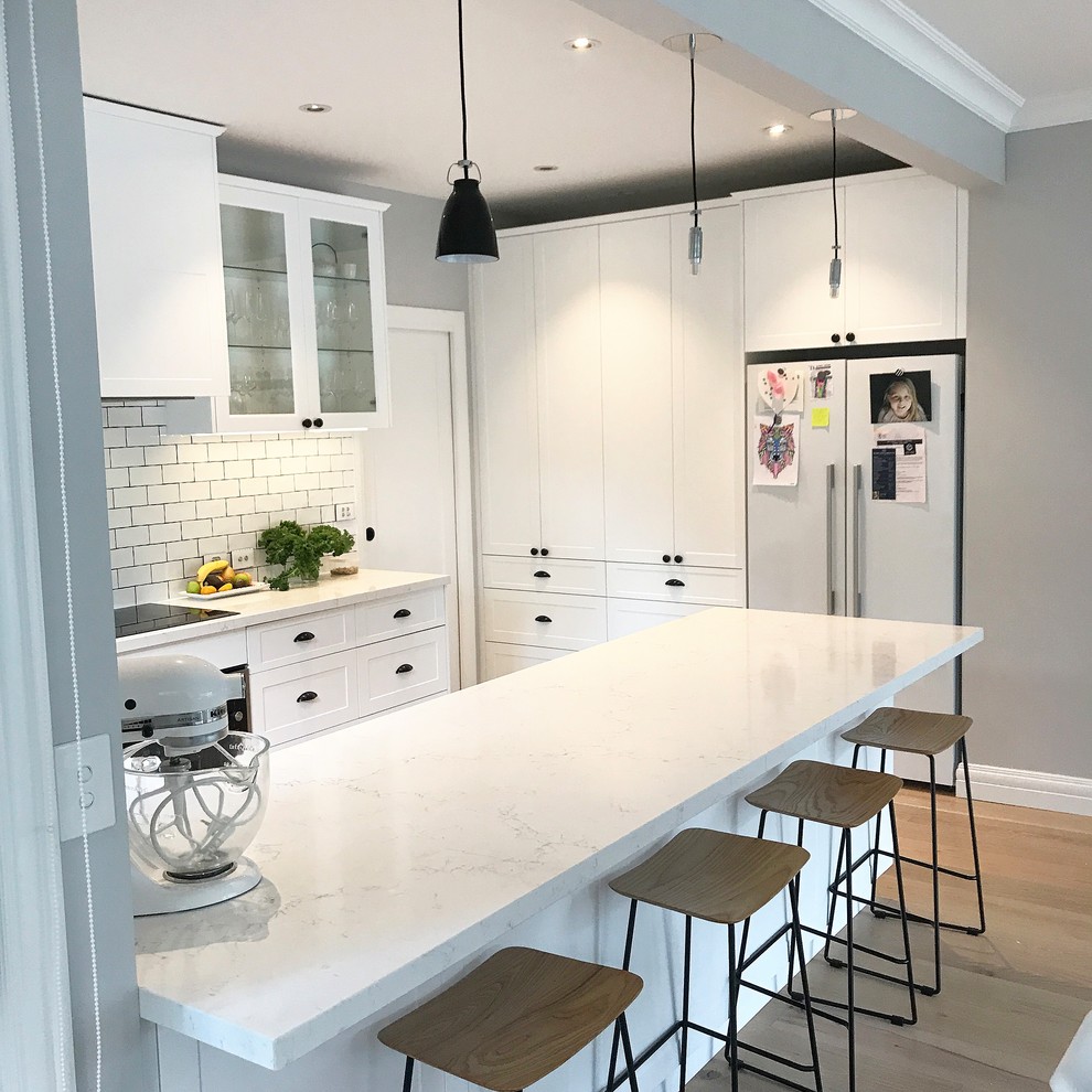 Mid-sized traditional u-shaped kitchen pantry in Melbourne with a drop-in sink, recessed-panel cabinets, white cabinets, quartz benchtops, white splashback, ceramic splashback, stainless steel appliances, light hardwood floors, with island and brown floor.