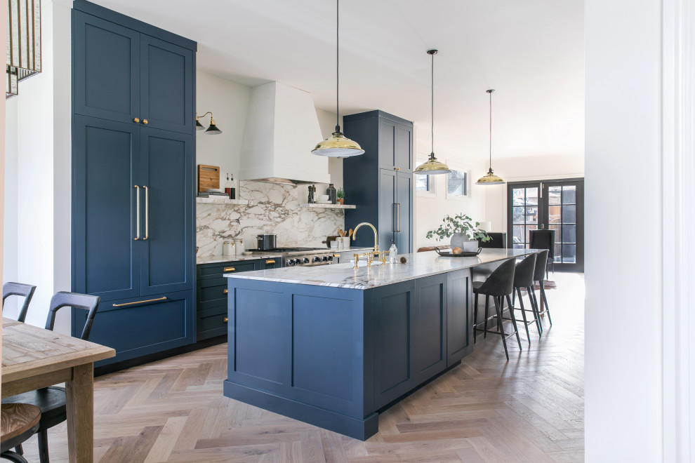 Photo of a mid-sized transitional galley open plan kitchen in Calgary with a double-bowl sink, shaker cabinets, blue cabinets, marble benchtops, multi-coloured splashback, marble splashback, panelled appliances, light hardwood floors, with island and multi-coloured benchtop.
