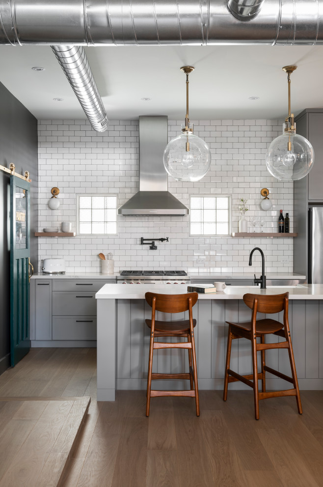 Inspiration for a mid-sized industrial l-shaped open plan kitchen in Toronto with a farmhouse sink, shaker cabinets, grey cabinets, quartz benchtops, white splashback, subway tile splashback, stainless steel appliances, light hardwood floors, with island, white benchtop and beige floor.