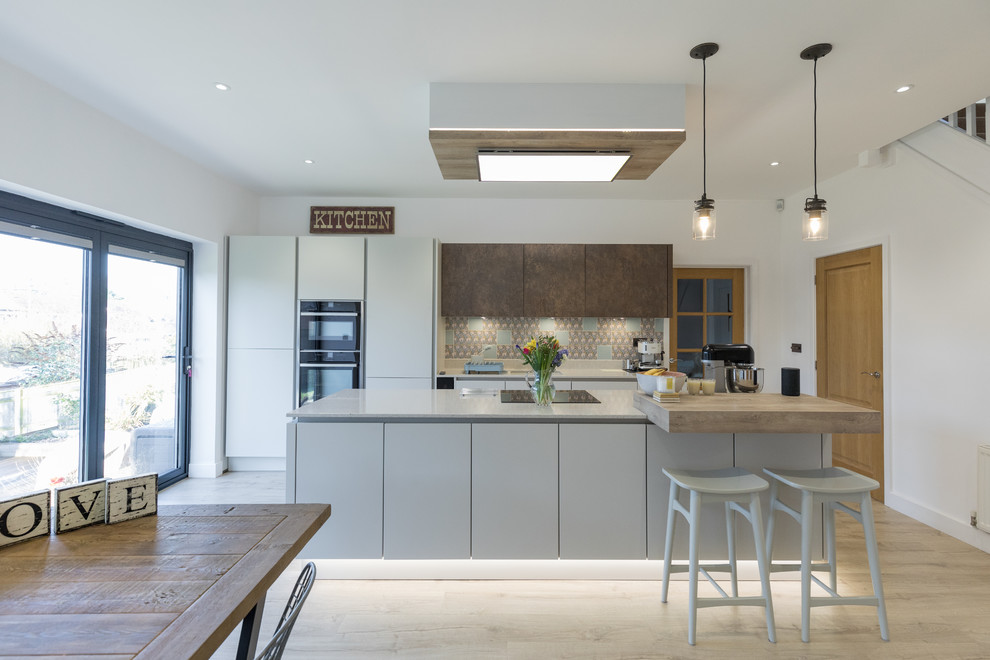 Mid-sized contemporary single-wall open plan kitchen in Hampshire with an undermount sink, flat-panel cabinets, white cabinets, quartzite benchtops, metallic splashback, panelled appliances, light hardwood floors and with island.