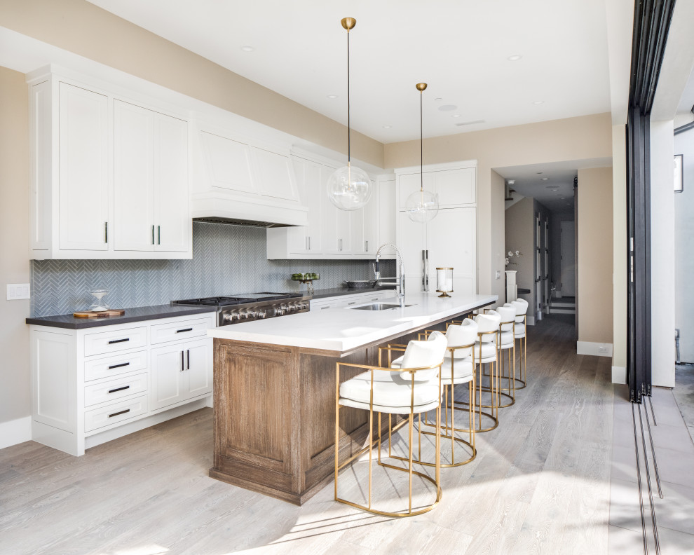Inspiration for a transitional l-shaped light wood floor and beige floor kitchen remodel in Orange County with an undermount sink, shaker cabinets, white cabinets, gray backsplash, mosaic tile backsplash, stainless steel appliances, an island and gray countertops