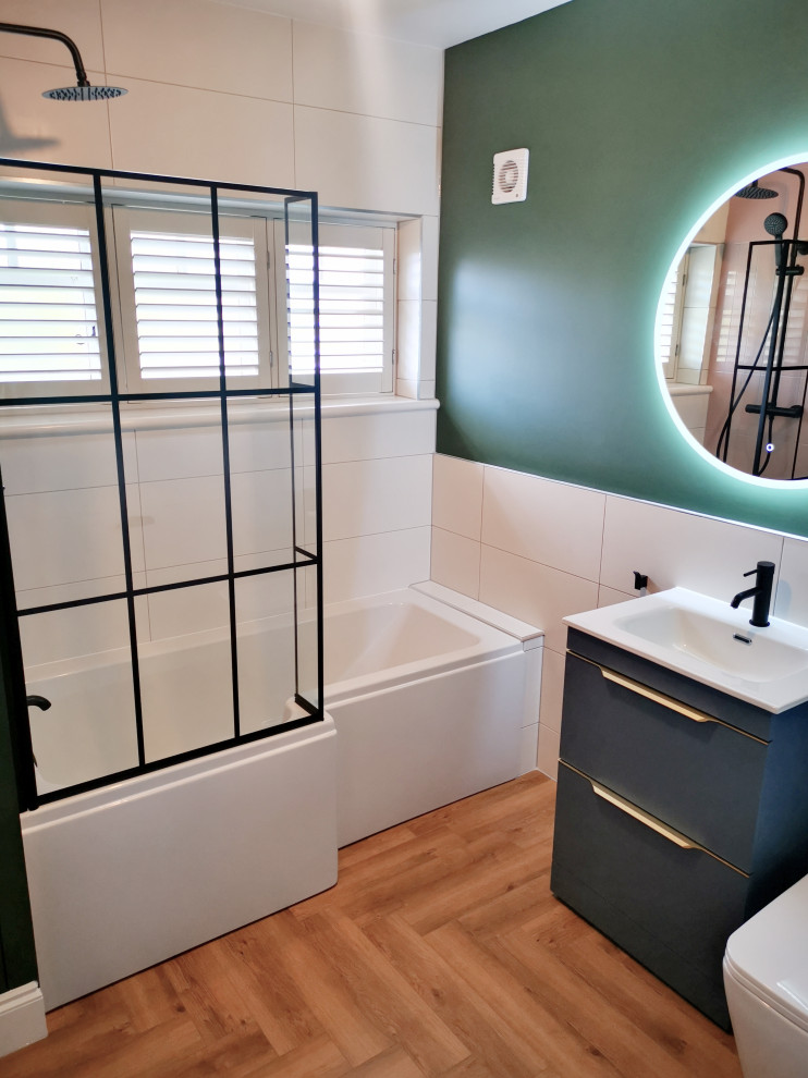 Photo of a mid-sized traditional kids bathroom in West Midlands with flat-panel cabinets, blue cabinets, a drop-in tub, a shower/bathtub combo, a one-piece toilet, pink tile, ceramic tile, green walls, vinyl floors, engineered quartz benchtops, white benchtops, a single vanity and a freestanding vanity.