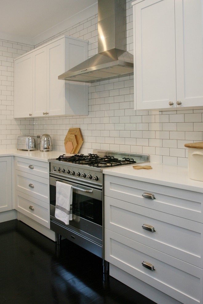Large traditional l-shaped open plan kitchen in Sydney with an integrated sink, shaker cabinets, white cabinets, white splashback, stainless steel appliances, with island, subway tile splashback, painted wood floors, black floor, white benchtop and solid surface benchtops.