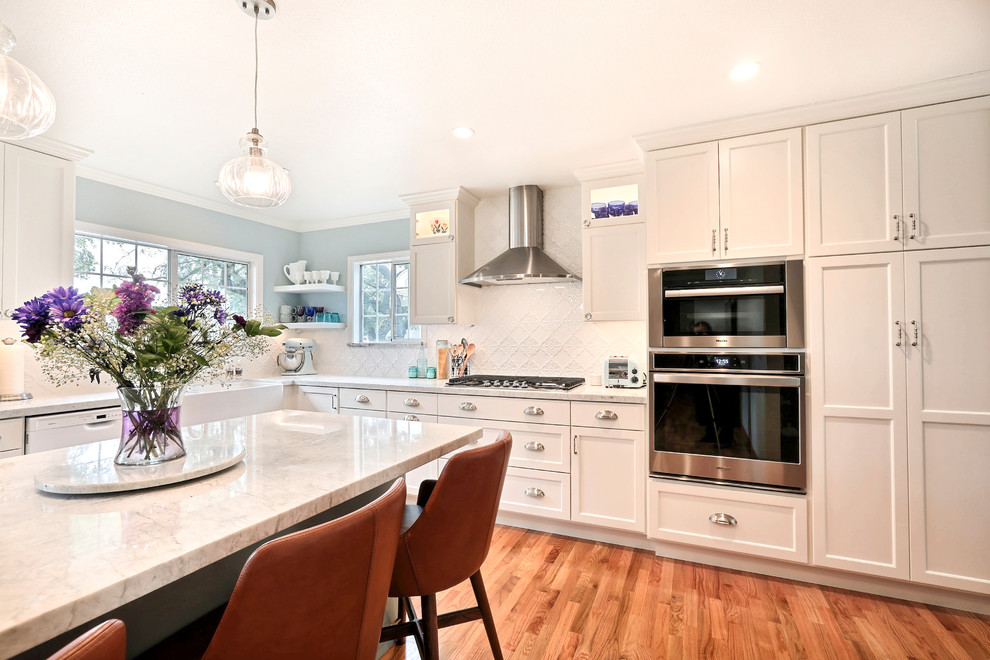 Mid-sized transitional l-shaped eat-in kitchen in Other with a farmhouse sink, shaker cabinets, white cabinets, quartzite benchtops, white splashback, ceramic splashback, stainless steel appliances, medium hardwood floors, with island and white benchtop.