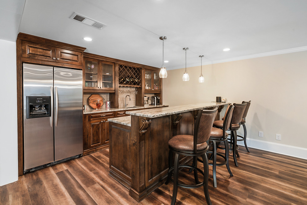 Large transitional single-wall seated home bar in Atlanta with a drop-in sink, glass-front cabinets, brown cabinets, granite benchtops, beige splashback, stone tile splashback, medium hardwood floors, brown floor and beige benchtop.