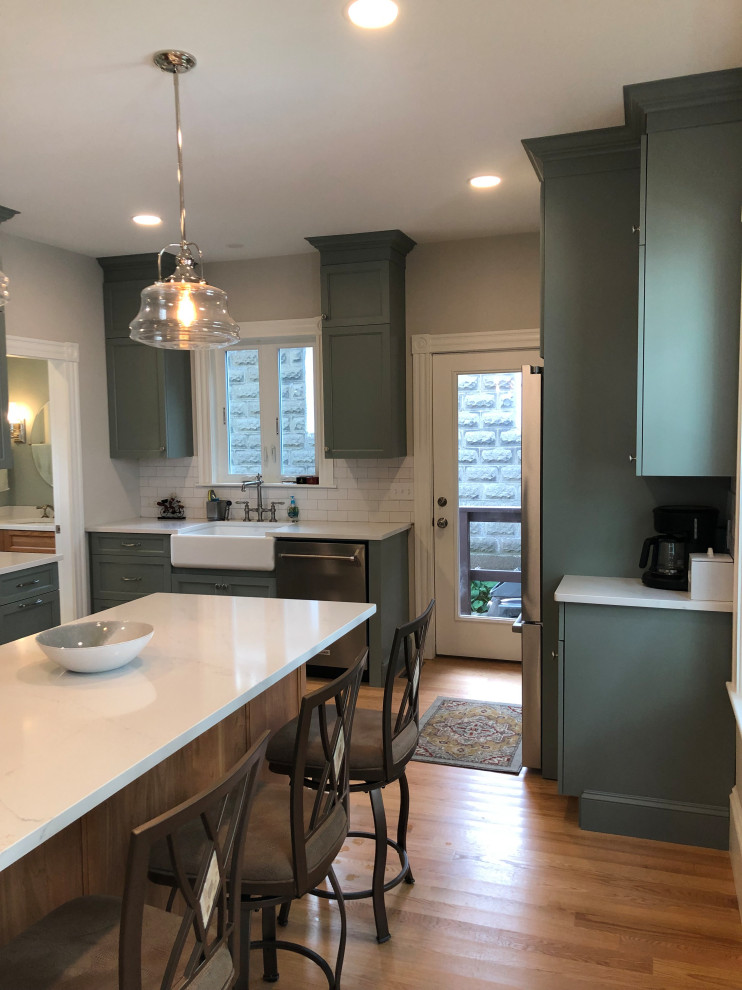 Example of a mid-sized classic u-shaped light wood floor and brown floor enclosed kitchen design in Boston with a farmhouse sink, flat-panel cabinets, green cabinets, quartz countertops, white backsplash, ceramic backsplash, stainless steel appliances, an island and white countertops