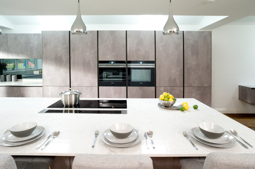 Photo of a contemporary eat-in kitchen in Essex with flat-panel cabinets, grey cabinets, quartz benchtops, grey splashback, mirror splashback, black appliances, medium hardwood floors, with island, brown floor and white benchtop.