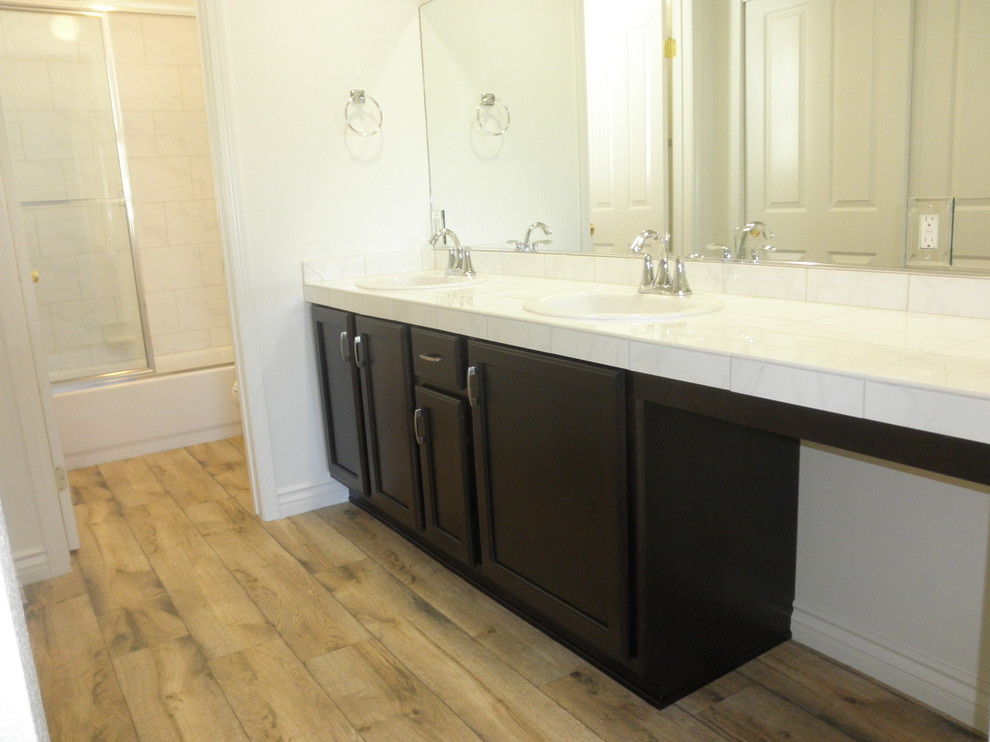 Inspiration for a transitional master beige tile and ceramic tile medium tone wood floor alcove bathtub remodel in Orange County with a drop-in sink, raised-panel cabinets, dark wood cabinets, tile countertops, a two-piece toilet and white walls