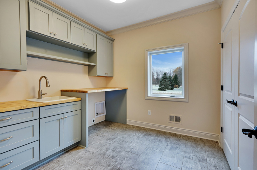 Inspiration for a large traditional single-wall dedicated laundry room in Detroit with a drop-in sink, shaker cabinets, grey cabinets, wood benchtops, beige walls, laminate floors, a side-by-side washer and dryer, grey floor and brown benchtop.