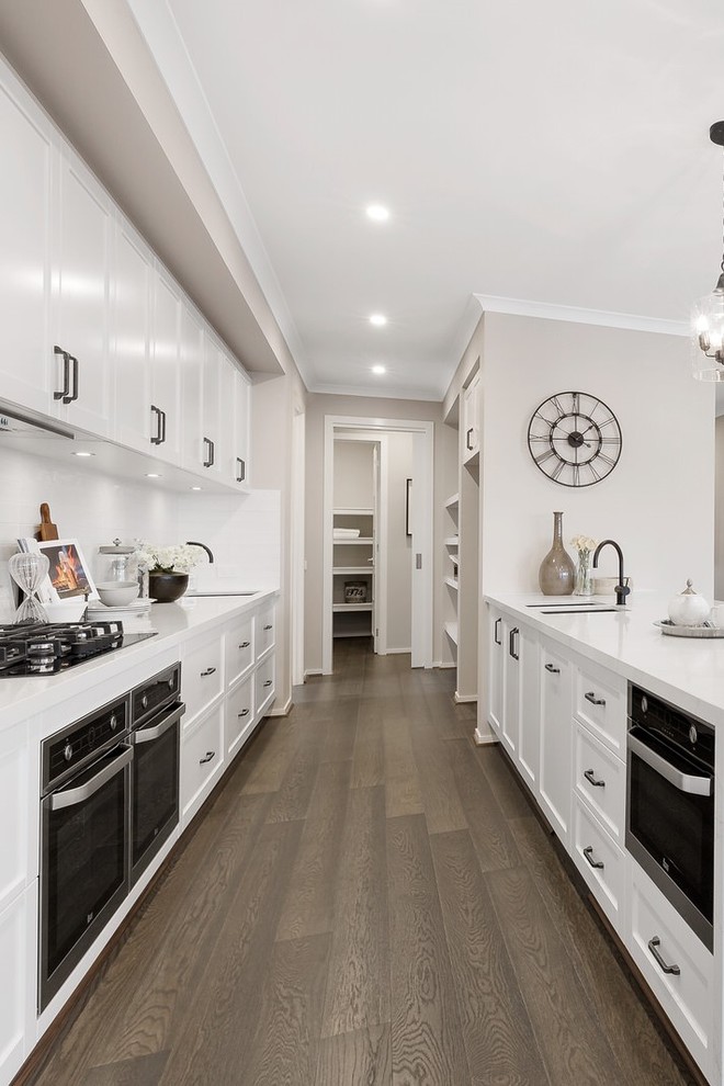 Photo of a beach style galley kitchen in Other with an undermount sink, recessed-panel cabinets, white cabinets, white splashback, black appliances, medium hardwood floors, brown floor and white benchtop.