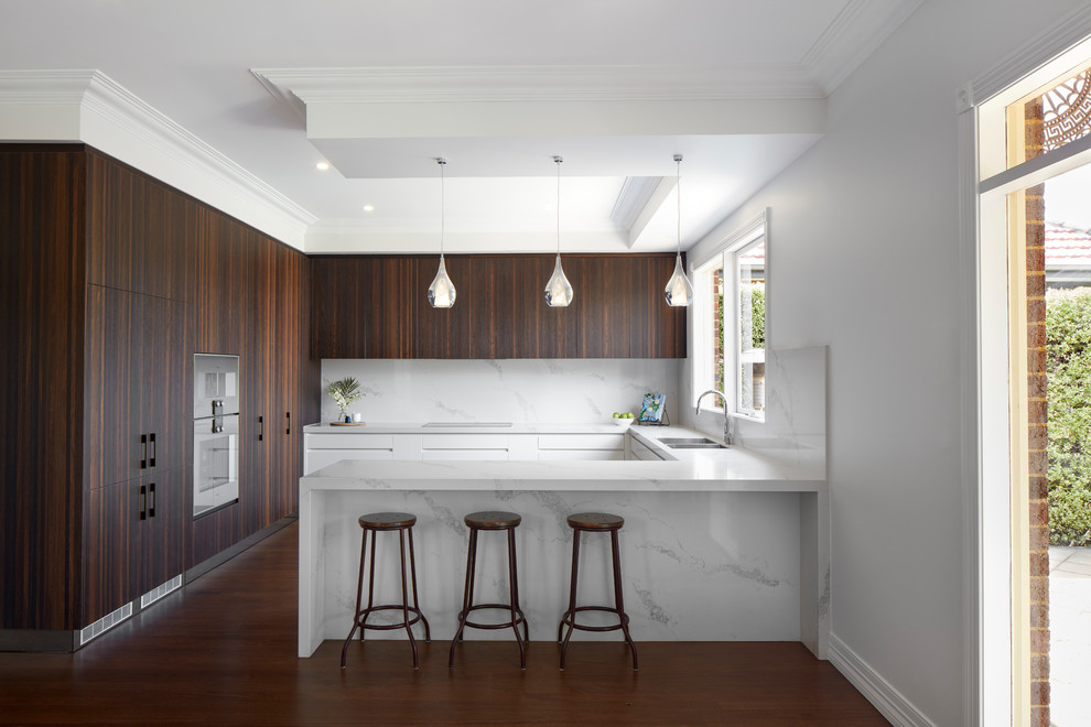 Photo of a large contemporary u-shaped kitchen in Melbourne with a double-bowl sink, flat-panel cabinets, dark wood cabinets, white splashback, stainless steel appliances, white benchtop, marble benchtops, marble splashback, light hardwood floors and with island.