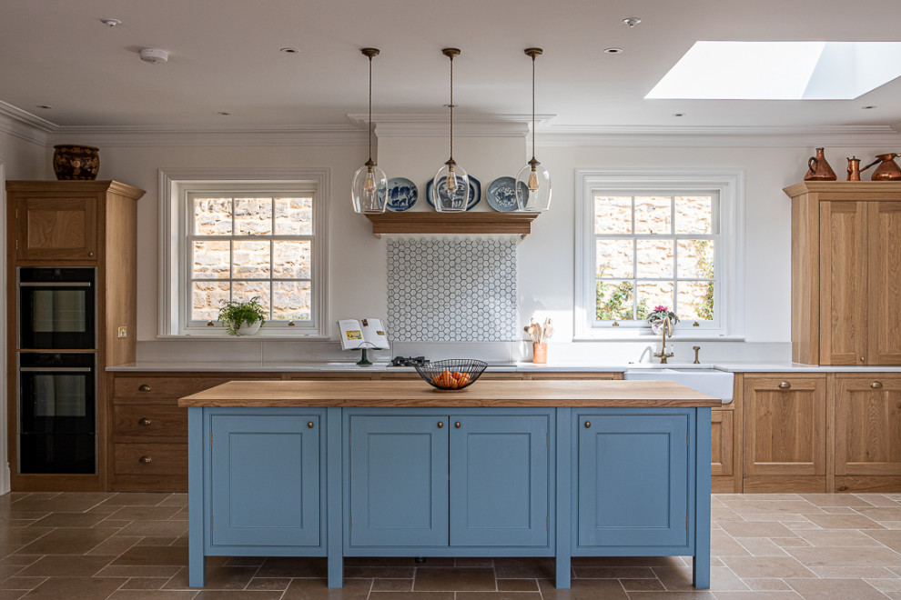 Inspiration for a large traditional kitchen in Dorset with a farmhouse sink, beaded inset cabinets, blue cabinets, wood benchtops, white splashback, black appliances, limestone floors, with island and beige floor.