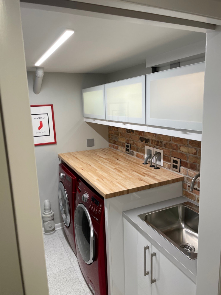 Medium sized retro galley separated utility room in Chicago with a built-in sink, glass-front cabinets, white cabinets, wood worktops, brick splashback, concrete flooring, a side by side washer and dryer and white floors.