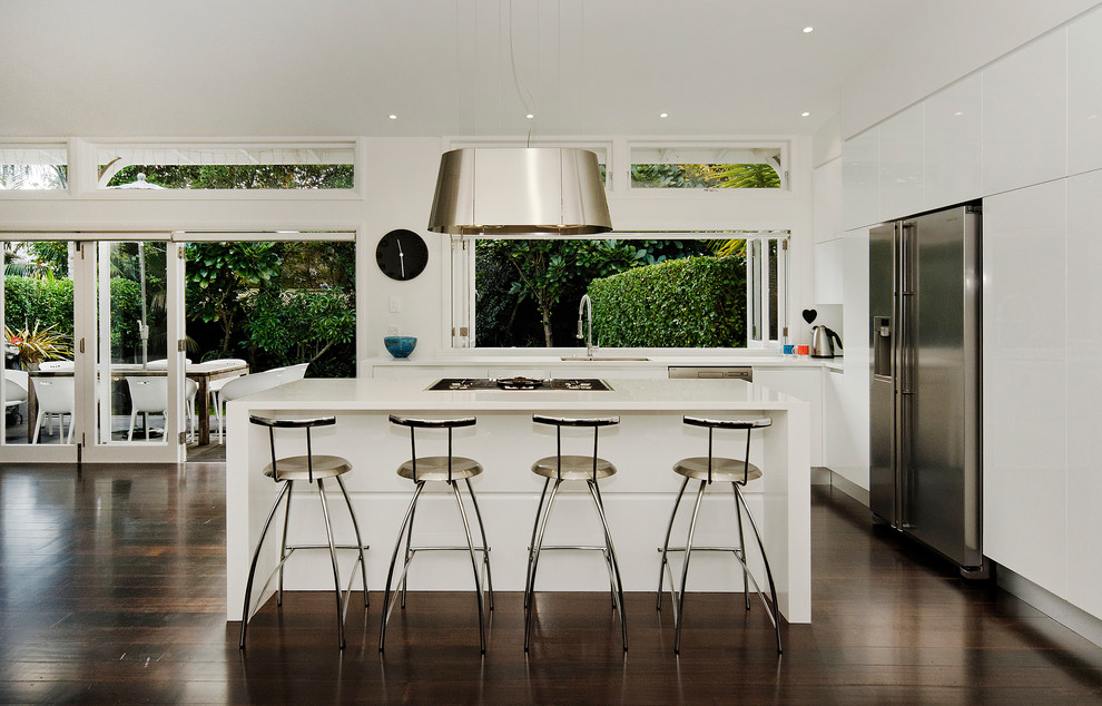 Mid-sized contemporary l-shaped eat-in kitchen in Auckland with stainless steel appliances, an undermount sink, flat-panel cabinets, white cabinets, solid surface benchtops, dark hardwood floors, with island and white splashback.