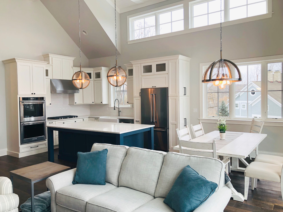 Large beach style l-shaped open plan kitchen in Chicago with a farmhouse sink, shaker cabinets, quartz benchtops, white splashback, subway tile splashback, stainless steel appliances, dark hardwood floors, with island, brown floor and white benchtop.