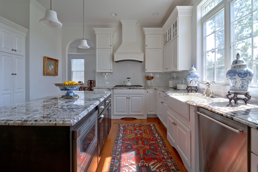 Inspiration for a traditional l-shaped kitchen in Boston with a farmhouse sink, raised-panel cabinets, white cabinets, white splashback, subway tile splashback, stainless steel appliances, medium hardwood floors and with island.