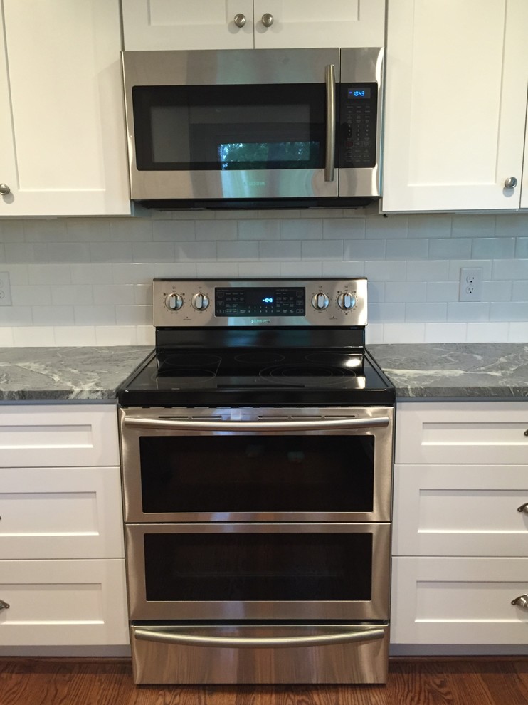 Example of a mid-sized transitional l-shaped medium tone wood floor enclosed kitchen design with an undermount sink, shaker cabinets, white cabinets, soapstone countertops, white backsplash, subway tile backsplash, stainless steel appliances and a peninsula