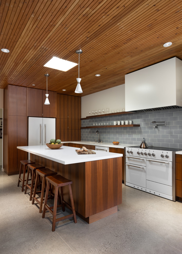 Photo of a midcentury galley eat-in kitchen in San Francisco with concrete floors, with island, exposed beam, flat-panel cabinets, medium wood cabinets, solid surface benchtops, blue splashback, ceramic splashback, white appliances, grey floor and white benchtop.