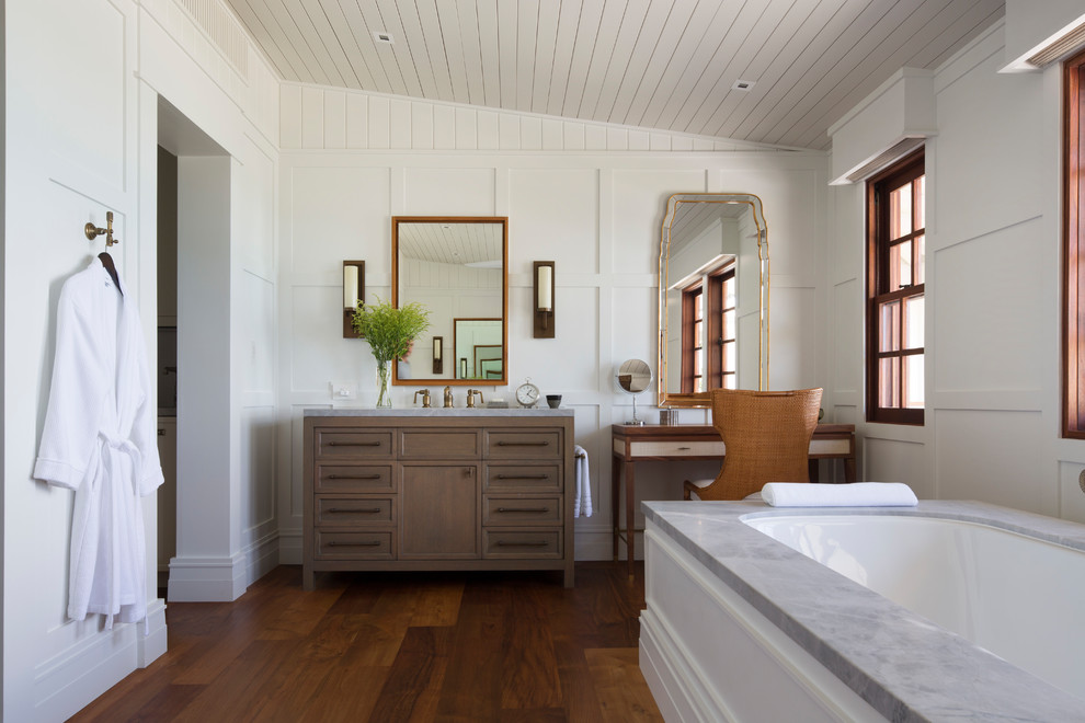 Design ideas for a tropical master bathroom in Hawaii with beaded inset cabinets, medium wood cabinets, a freestanding tub, a one-piece toilet, gray tile, white walls, medium hardwood floors, an undermount sink, marble benchtops, brown floor, a hinged shower door and grey benchtops.