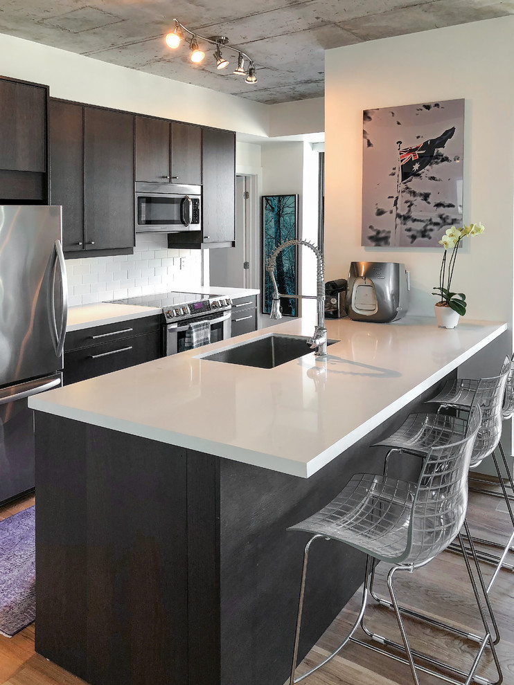 This is an example of a mid-sized modern galley open plan kitchen in Toronto with an undermount sink, flat-panel cabinets, dark wood cabinets, quartz benchtops, white splashback, ceramic splashback, stainless steel appliances, medium hardwood floors, a peninsula, brown floor and white benchtop.