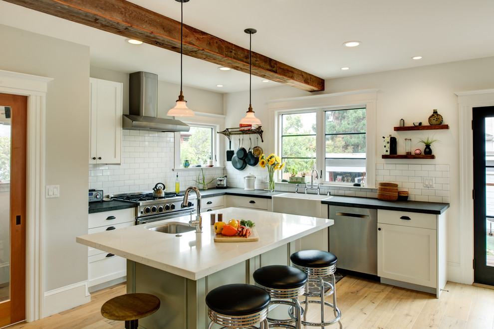 Inspiration for a mid-sized eclectic l-shaped open plan kitchen in San Francisco with a farmhouse sink, shaker cabinets, white cabinets, soapstone benchtops, white splashback, subway tile splashback, stainless steel appliances, light hardwood floors, with island and brown floor.