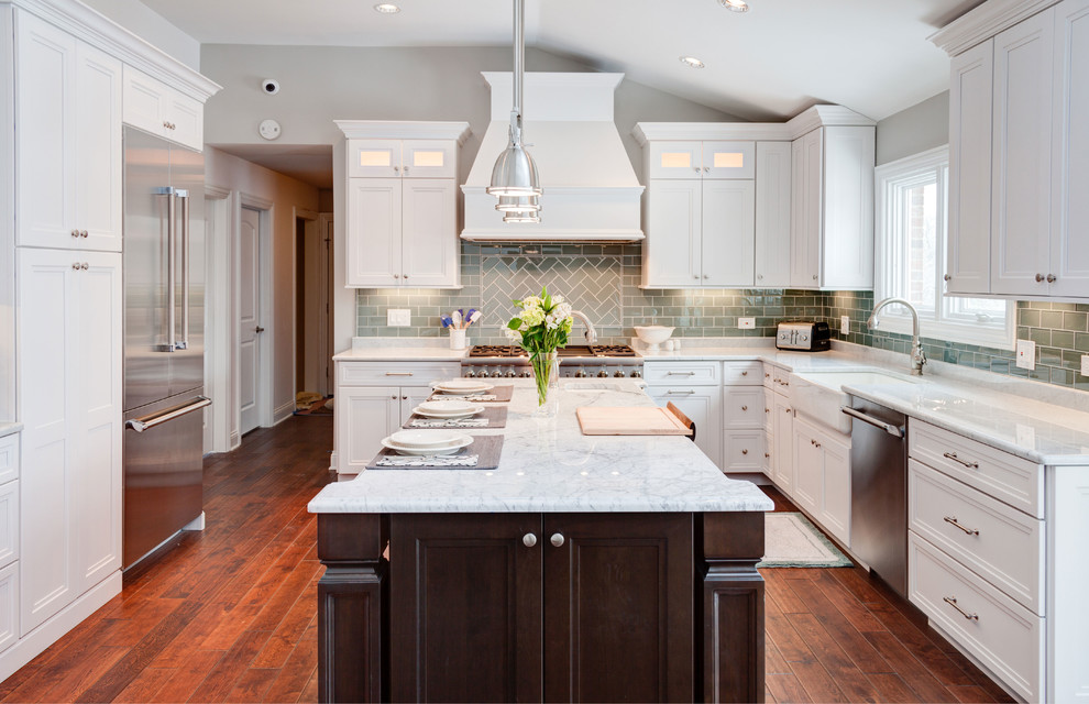 This is an example of a transitional u-shaped eat-in kitchen in Chicago with a farmhouse sink, flat-panel cabinets, white cabinets, marble benchtops, glass tile splashback, stainless steel appliances, grey splashback, medium hardwood floors, with island and brown floor.
