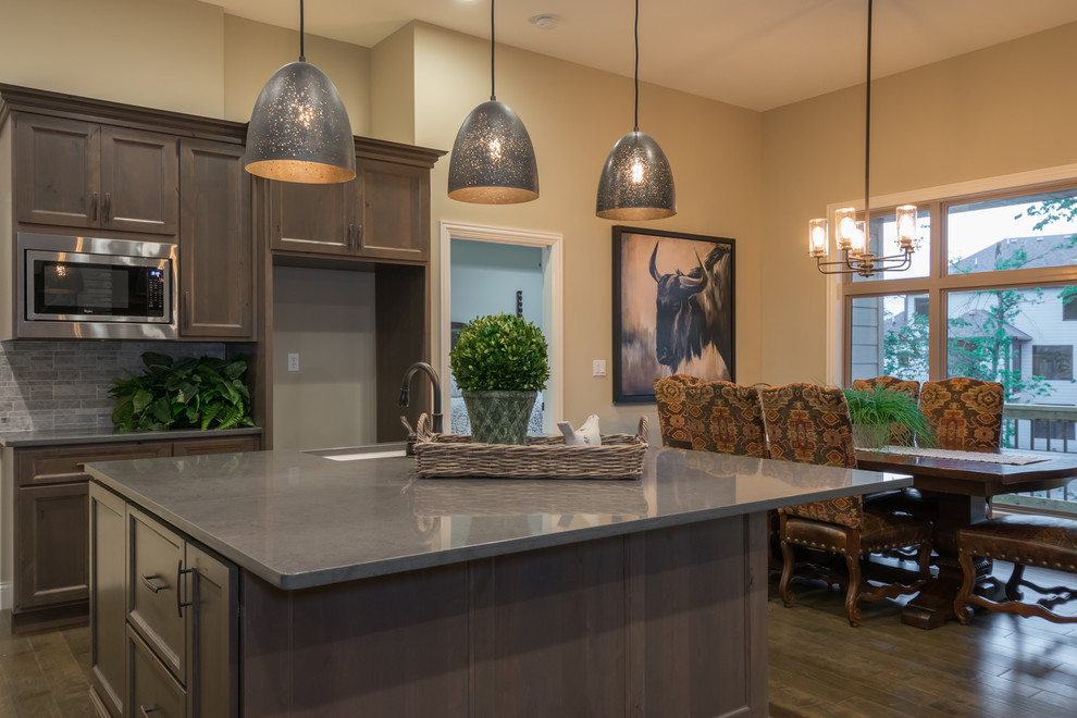 Inspiration for a large rustic galley medium tone wood floor and brown floor open concept kitchen remodel with a single-bowl sink, shaker cabinets, brown cabinets, quartz countertops, gray backsplash, stone tile backsplash, stainless steel appliances and an island