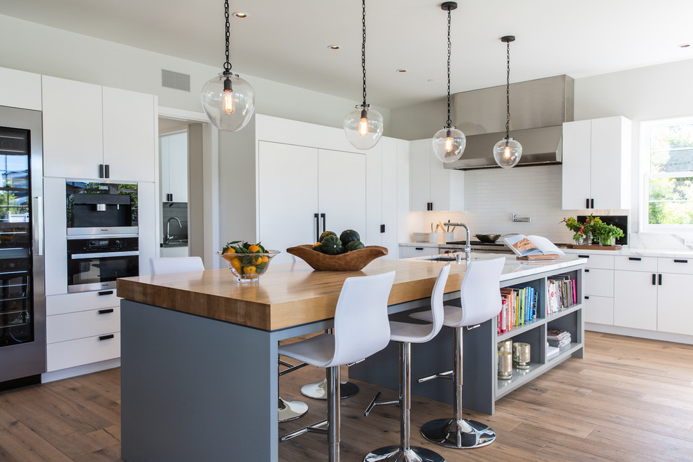 Photo of a contemporary kitchen in Los Angeles with an undermount sink, flat-panel cabinets, white cabinets, wood benchtops, white splashback, stainless steel appliances, light hardwood floors and with island.