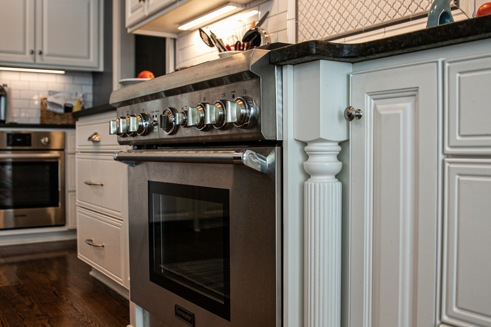 Example of a large transitional l-shaped medium tone wood floor and brown floor open concept kitchen design in Charlotte with a farmhouse sink, white cabinets, granite countertops, white backsplash, stainless steel appliances, an island, raised-panel cabinets, porcelain backsplash and white countertops
