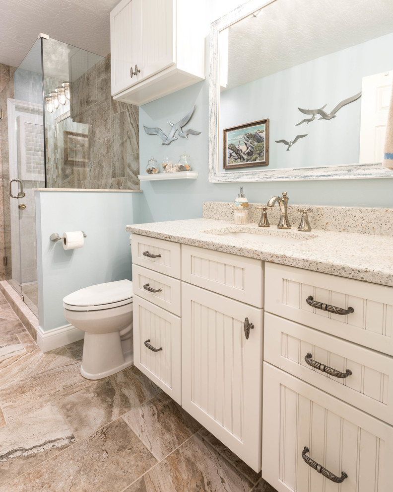 This is an example of a large beach style 3/4 bathroom in Other with beaded inset cabinets, white cabinets, a corner shower, beige tile, porcelain tile, blue walls, porcelain floors, an undermount sink, recycled glass benchtops, beige floor and a hinged shower door.