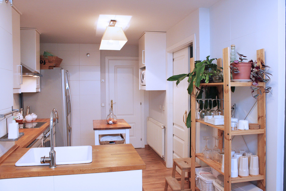 Ejemplo de cocina lineal escandinava abierta con fregadero de un seno, armarios con paneles lisos, puertas de armario blancas, encimera de madera, salpicadero blanco, salpicadero de azulejos de cerámica, electrodomésticos de acero inoxidable y suelo de madera en tonos medios