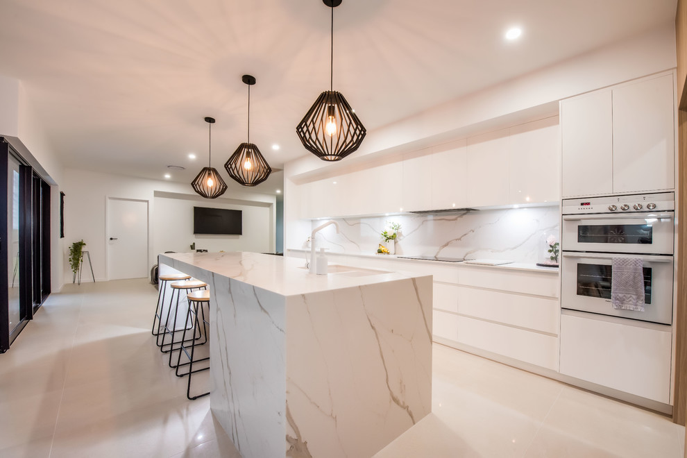 This is an example of a large contemporary galley open plan kitchen in Other with flat-panel cabinets, white splashback, marble splashback, white appliances, white benchtop, with island, an undermount sink, light wood cabinets, marble benchtops, ceramic floors and white floor.