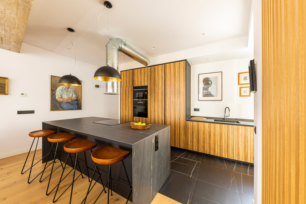 This is an example of an industrial galley kitchen in Madrid with an undermount sink, flat-panel cabinets, medium wood cabinets, black appliances, with island, black floor and grey benchtop.