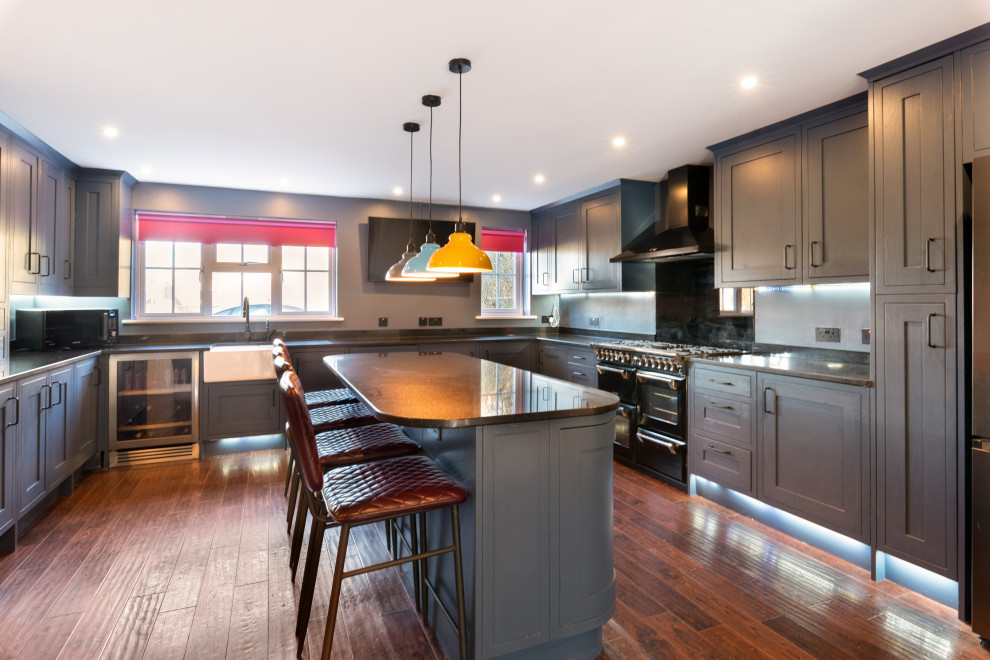 Example of a large trendy u-shaped dark wood floor and brown floor open concept kitchen design in Kent with a farmhouse sink, blue cabinets, quartzite countertops, black backsplash, glass sheet backsplash, paneled appliances, an island, black countertops and shaker cabinets