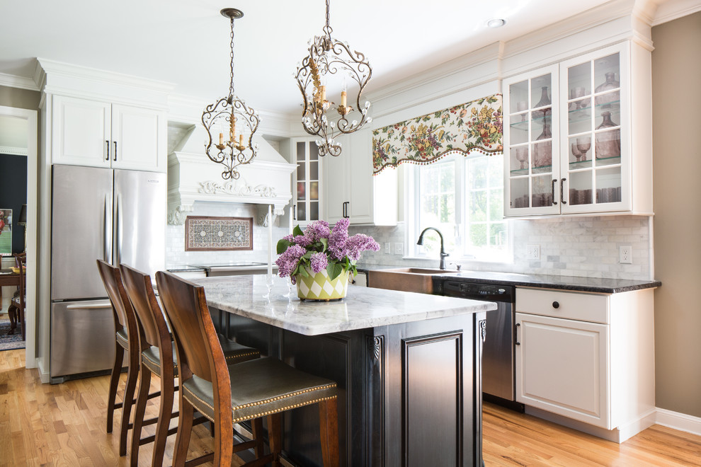 Photo of a traditional l-shaped kitchen in Boston with a farmhouse sink, raised-panel cabinets, white cabinets, grey splashback, stone tile splashback, stainless steel appliances, light hardwood floors and with island.