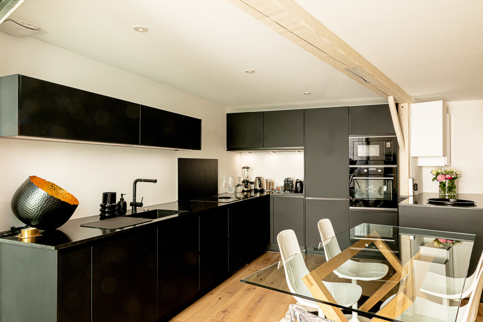 Expansive contemporary l-shaped open plan kitchen in Other with a submerged sink, granite worktops, black splashback, black appliances, no island, brown floors, black worktops and exposed beams.
