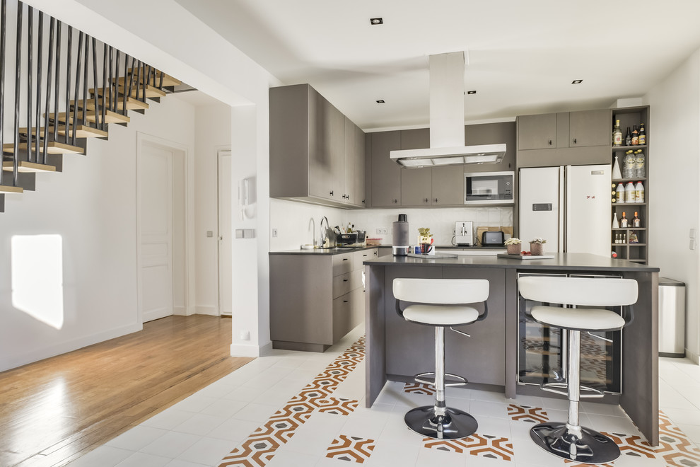 This is an example of a contemporary l-shaped kitchen in Paris with flat-panel cabinets, grey cabinets, white splashback, stainless steel appliances, with island, multi-coloured floor and grey benchtop.