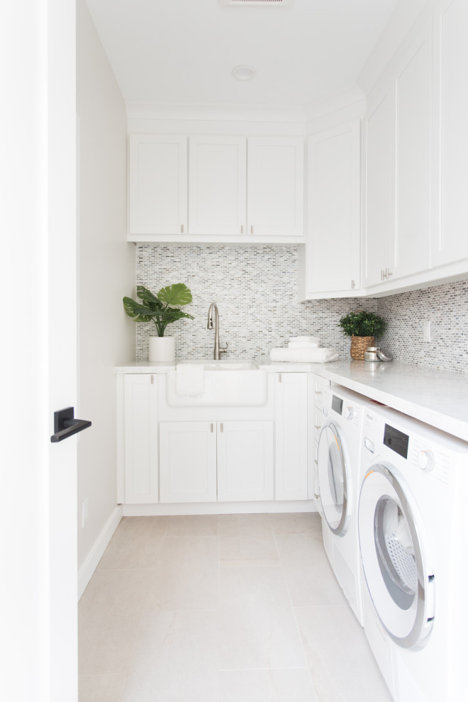 Photo of a mid-sized transitional l-shaped dedicated laundry room in Santa Barbara with a farmhouse sink, shaker cabinets, white cabinets, white walls, porcelain floors, a side-by-side washer and dryer, beige floor, white benchtop and marble benchtops.