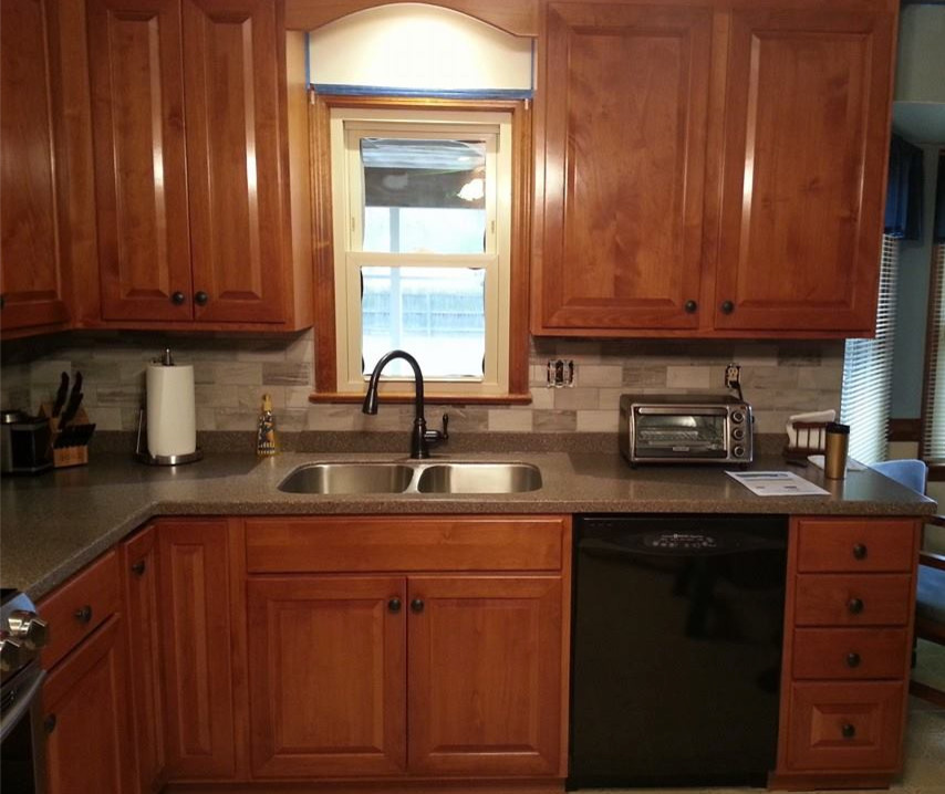 L-shaped eat-in kitchen photo with a drop-in sink, raised-panel cabinets, medium tone wood cabinets, laminate countertops, gray backsplash, ceramic backsplash and black appliances