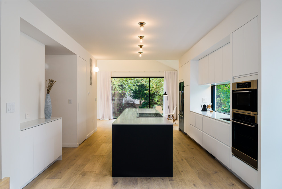 Mid-sized scandinavian galley open plan kitchen in Philadelphia with an undermount sink, flat-panel cabinets, white cabinets, solid surface benchtops, window splashback, black appliances, light hardwood floors, with island, beige floor and white benchtop.
